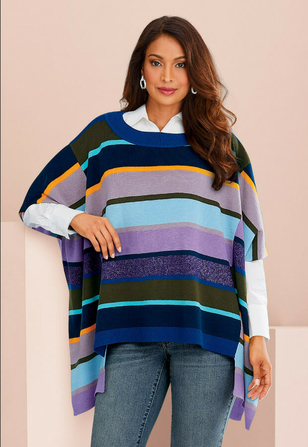 A woman in a colorful striped poncho poses against a soft pink background, wearing a white shirt and stylish earrings.
