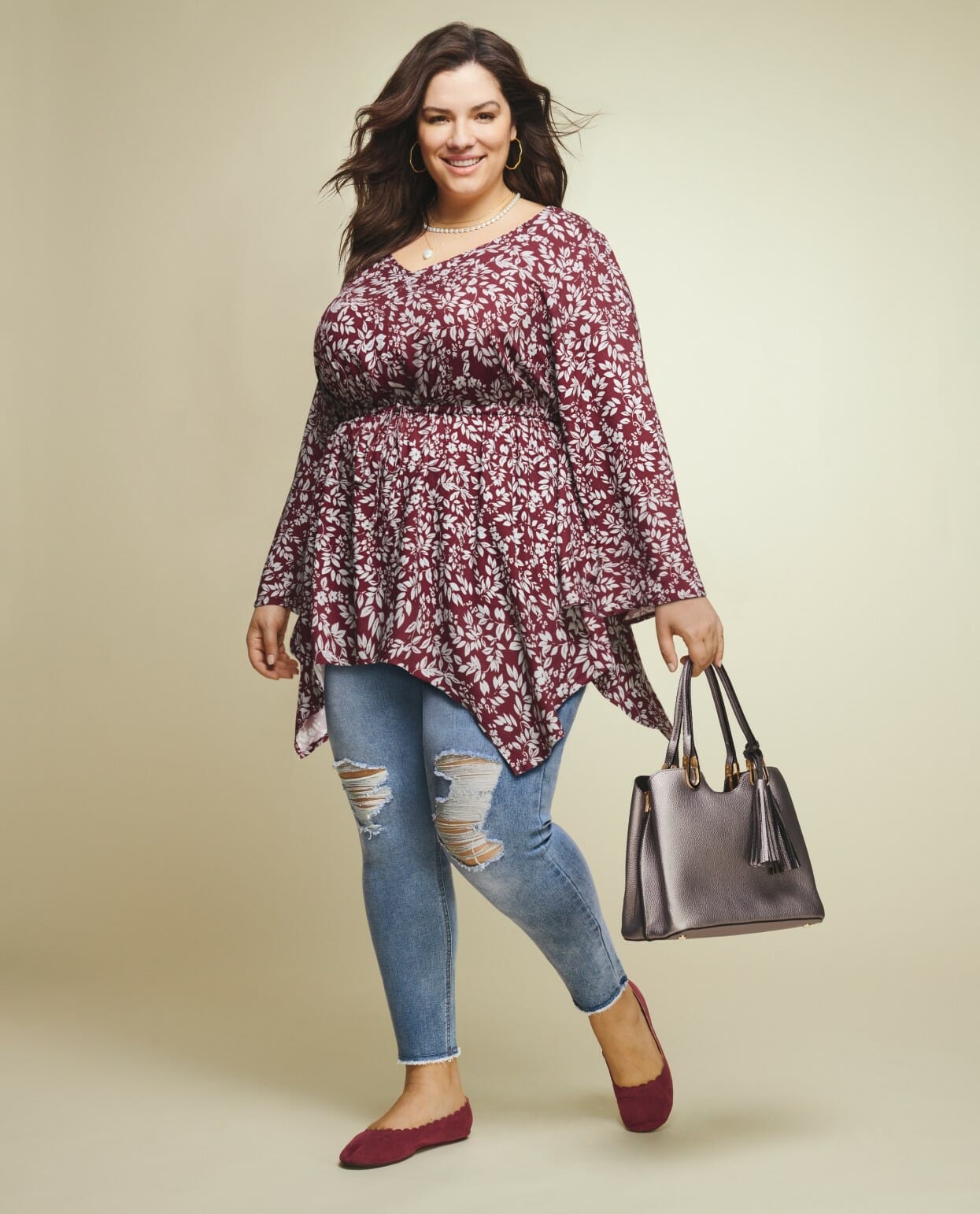 Plus size model wearing a burgundy floral blouse with distressed jeans, carrying a silver purse.