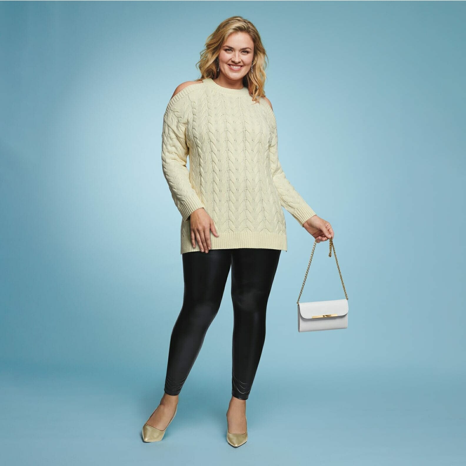 Plus size model wearing cream sweater with leather leggings holding a white purse.