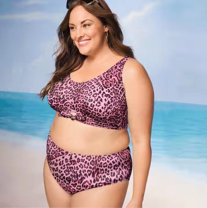 Smiling woman in a pink leopard print bikini stands by the beach, with ocean waves and a clear sky in the background.