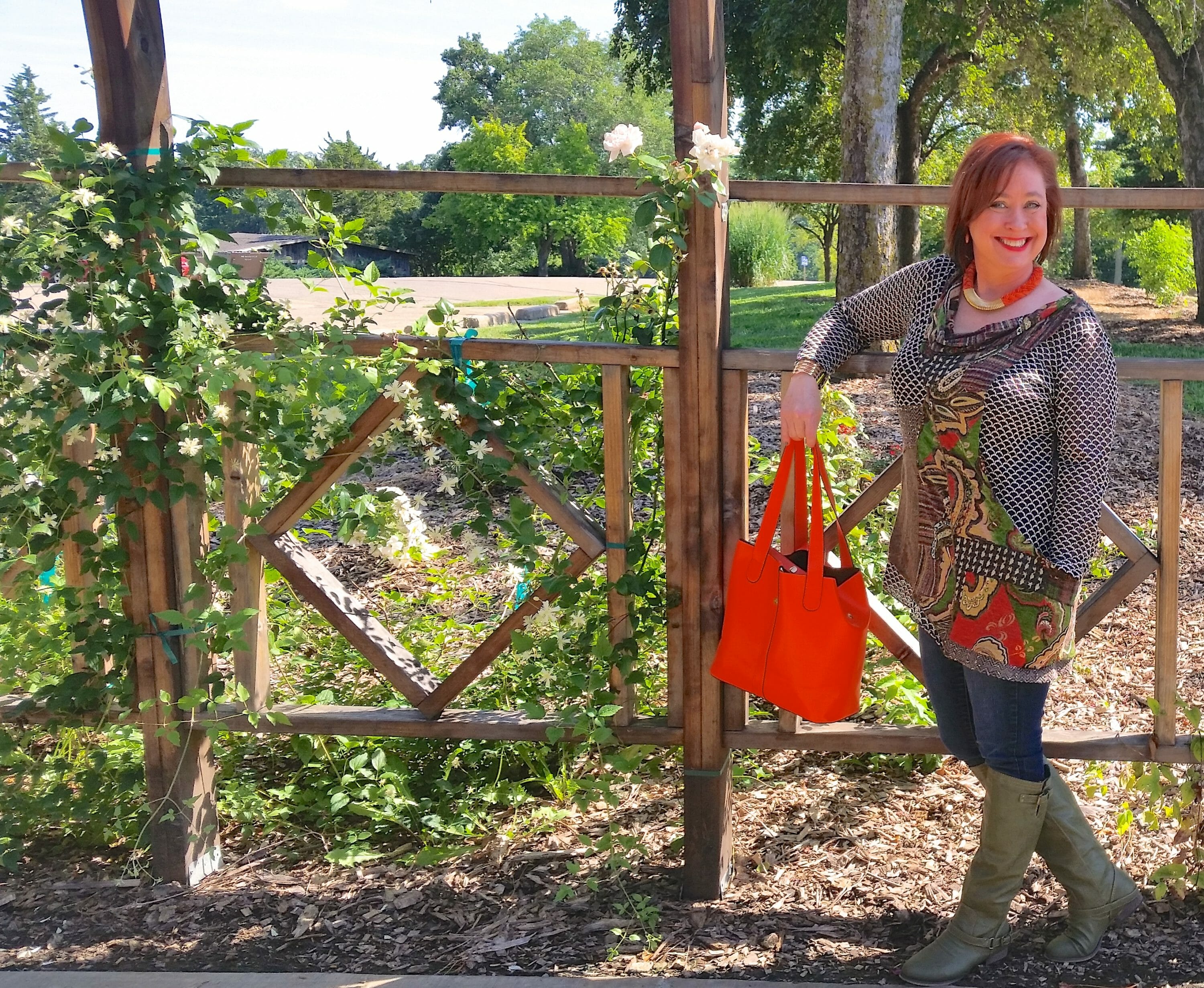 woman standing in front of gate in a top with orange accents and a orange handbag