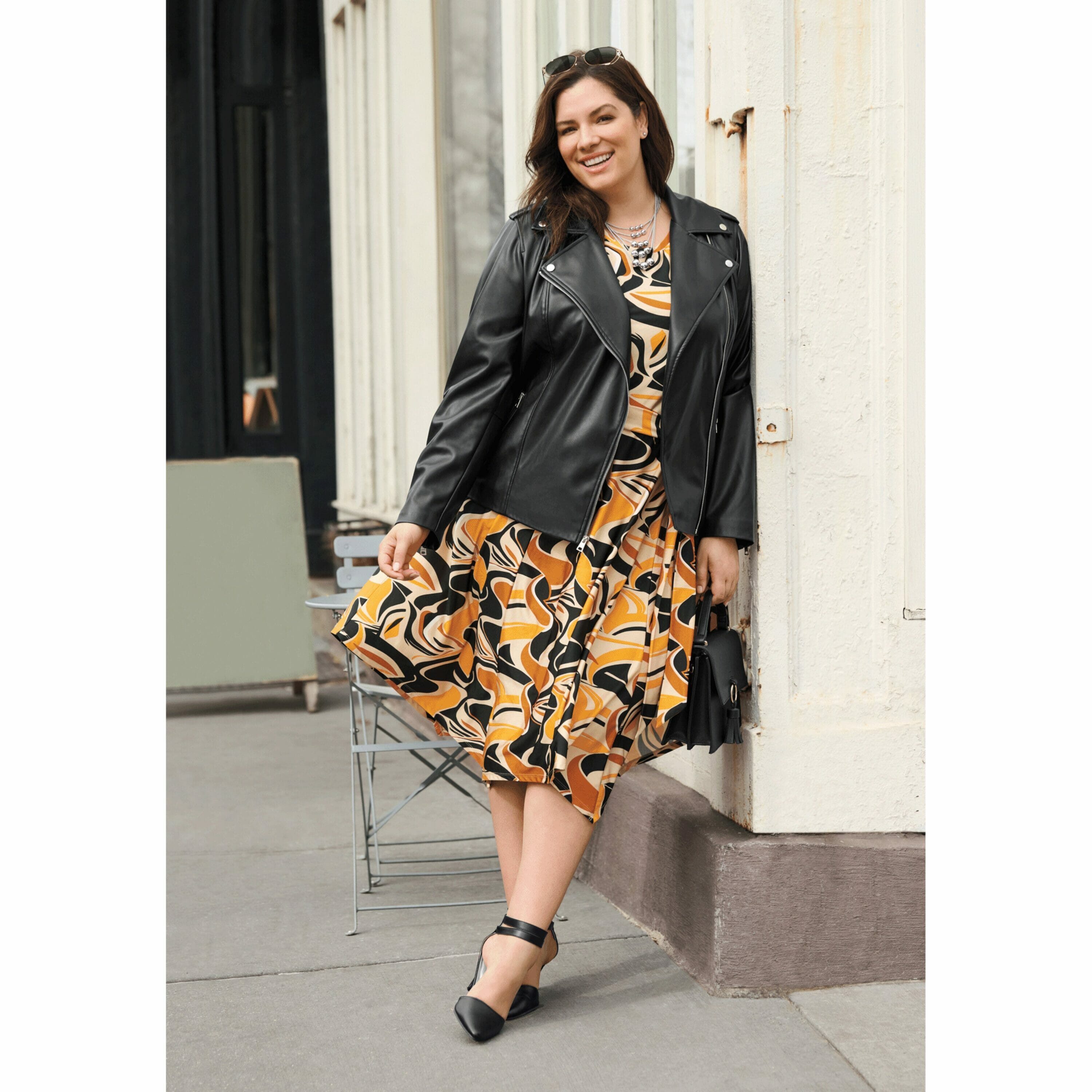 plus size model wearing a printed yellow and orange dress with a leather jacket.