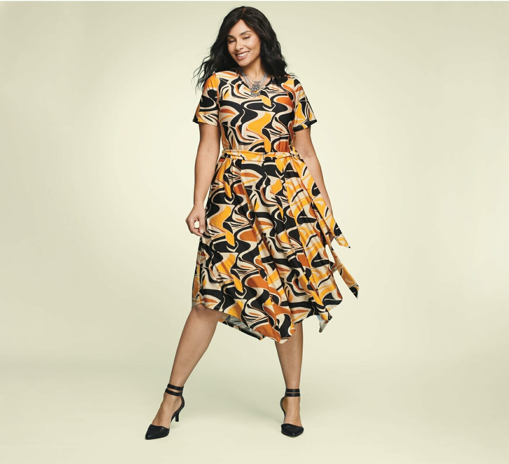 plus size model wearing a printed orange and yellow dress with black pumps.