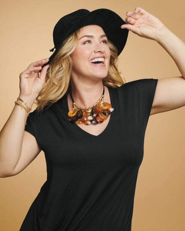 plus size model wearing a black t-shirt, gaudy necklace, with a black hat and a layered teardrop necklace.